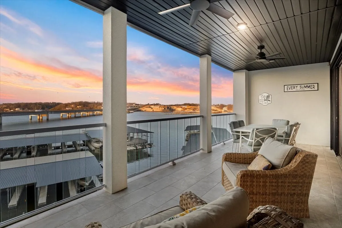 The image captures an inviting outdoor seating area overlooking the expansive waters of Lake of the Ozarks during a vibrant sunset. The scene radiates tranquility, with comfortable seating ideal for relaxing or socializing, and a decorative sign that reads 'EVERY SUMMER,' reinforcing the property's seasonal appeal. The view stretches across the main channel, with rich sunset hues painting the sky, perfectly illustrating lakeside serenity.
