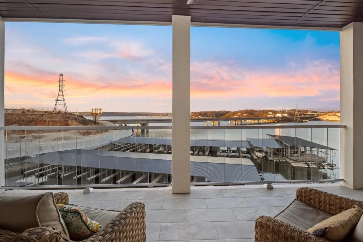 A serene water view from a balcony furnished with cozy seating. The sunset sky casts warm hues over the calm lake, creating a peaceful and inviting space for morning coffee or evening relaxation.