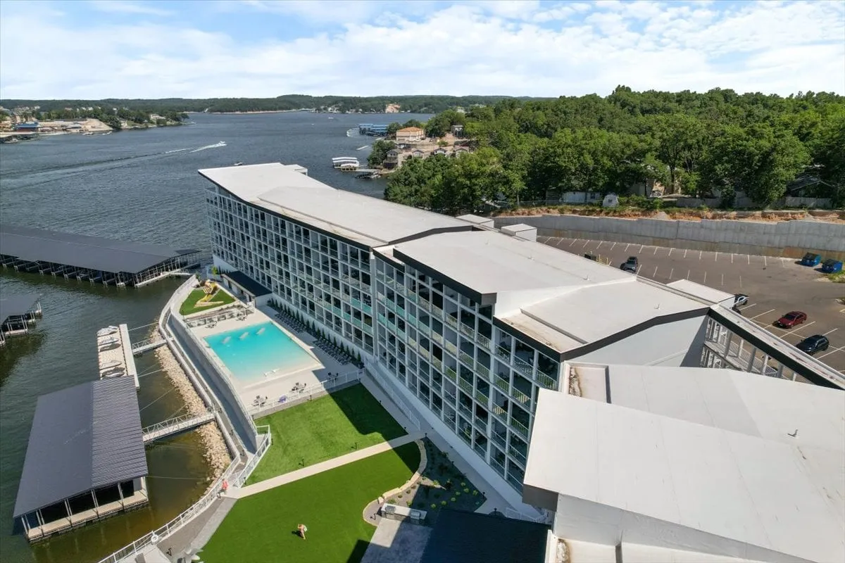 This image captures a vibrant exterior scene featuring a swimming pool, expansive waterfront views, the modern building structure, and a lush green lawn. The composition includes people enjoying the pool and relaxing by the water, highlighting both active and tranquil moments.