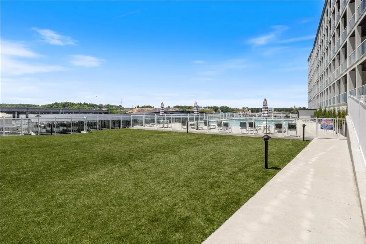 An exterior shot with an expansive green lawn, a sitting area shaded by umbrellas, the swimming pool in the background, and the modern building facade. The inviting outdoor space is ideal for gatherings, lawn games, yoga, or simply relaxing after a day of exploration.