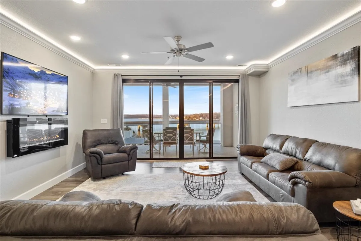 A luxurious living room with a modern fireplace, comfortable seating arrangement, decorative artwork, and expansive glass sliding doors. The doors open directly toward the balcony, offering unobstructed views of the lake and inviting natural light into the space. The ambiance is cozy, with a blend of relaxation and sophistication.