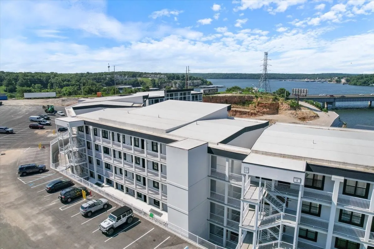 An inviting view of the building structure set beside a nearby water body, with a green backdrop and a visible parking lot. The scene conveys easy access to water sports, fishing, picnicking, and exploration of nearby trails.