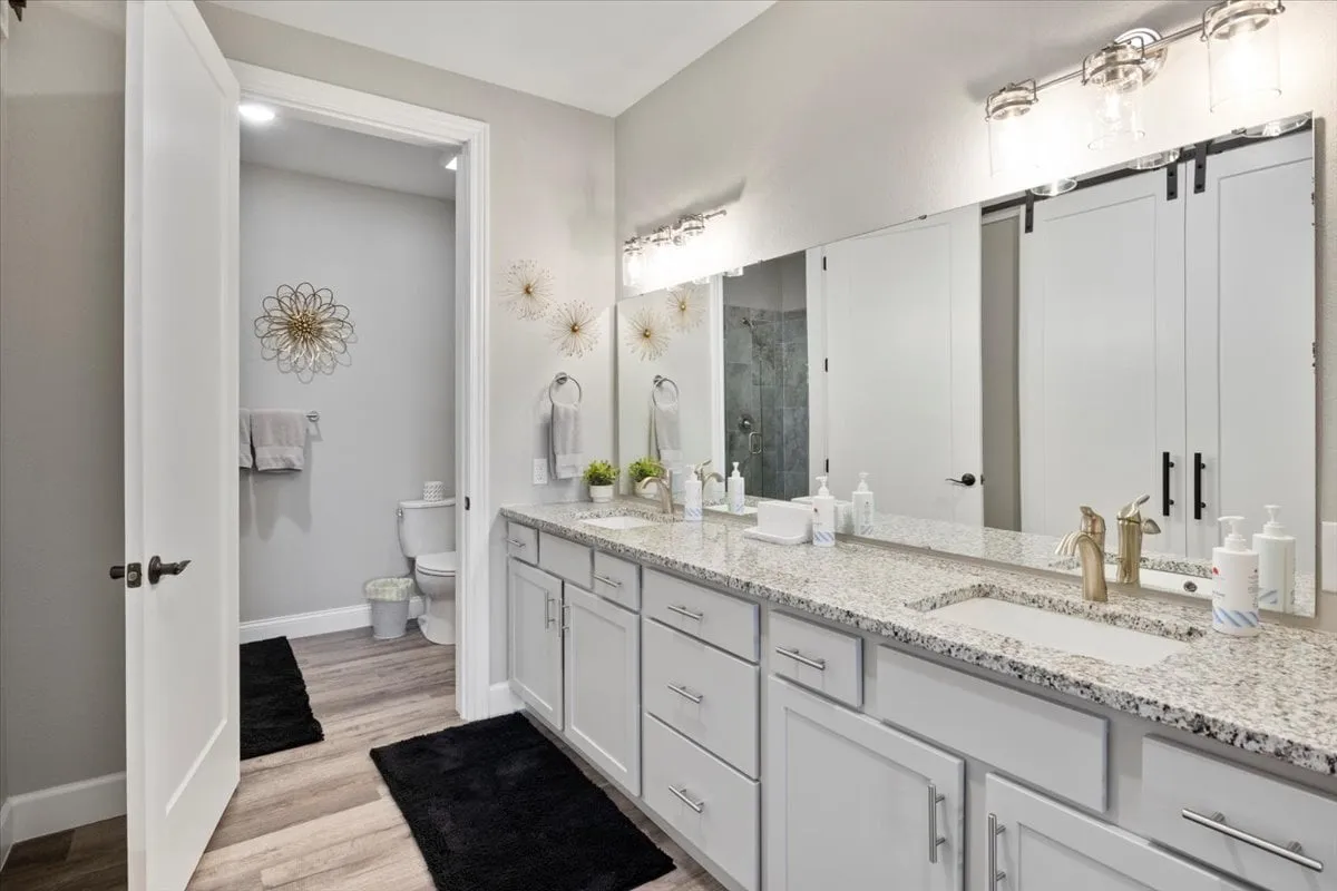 The image features a modern ensuite bathroom with a double vanity, granite countertop, and large mirrors. Decorative wall art and a soothing color palette evoke a spa-like, serene mood. The space is bright, clean, and perfect for rejuvenation and self-care.