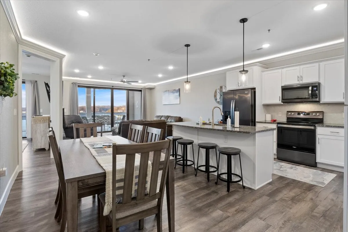 A warm, welcoming open-plan space featuring a dining table, kitchen island, and a spacious living area—all oriented toward oversized windows that look out to the balcony and lake. The layout encourages easy flow and social interaction, with sunlight streaming in and an inviting atmosphere for family dinners or entertaining guests.